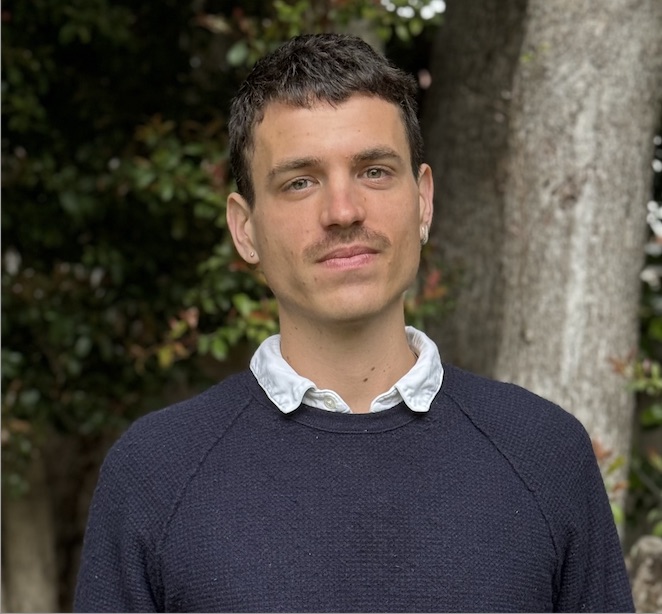 A young man with short, brown hair, wearing a sweater, standing in front of trees.
